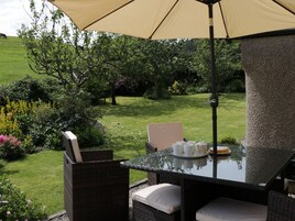 Un patio exterior con mesa de vidrio, sillas de mimbre y un paraguas color beige.