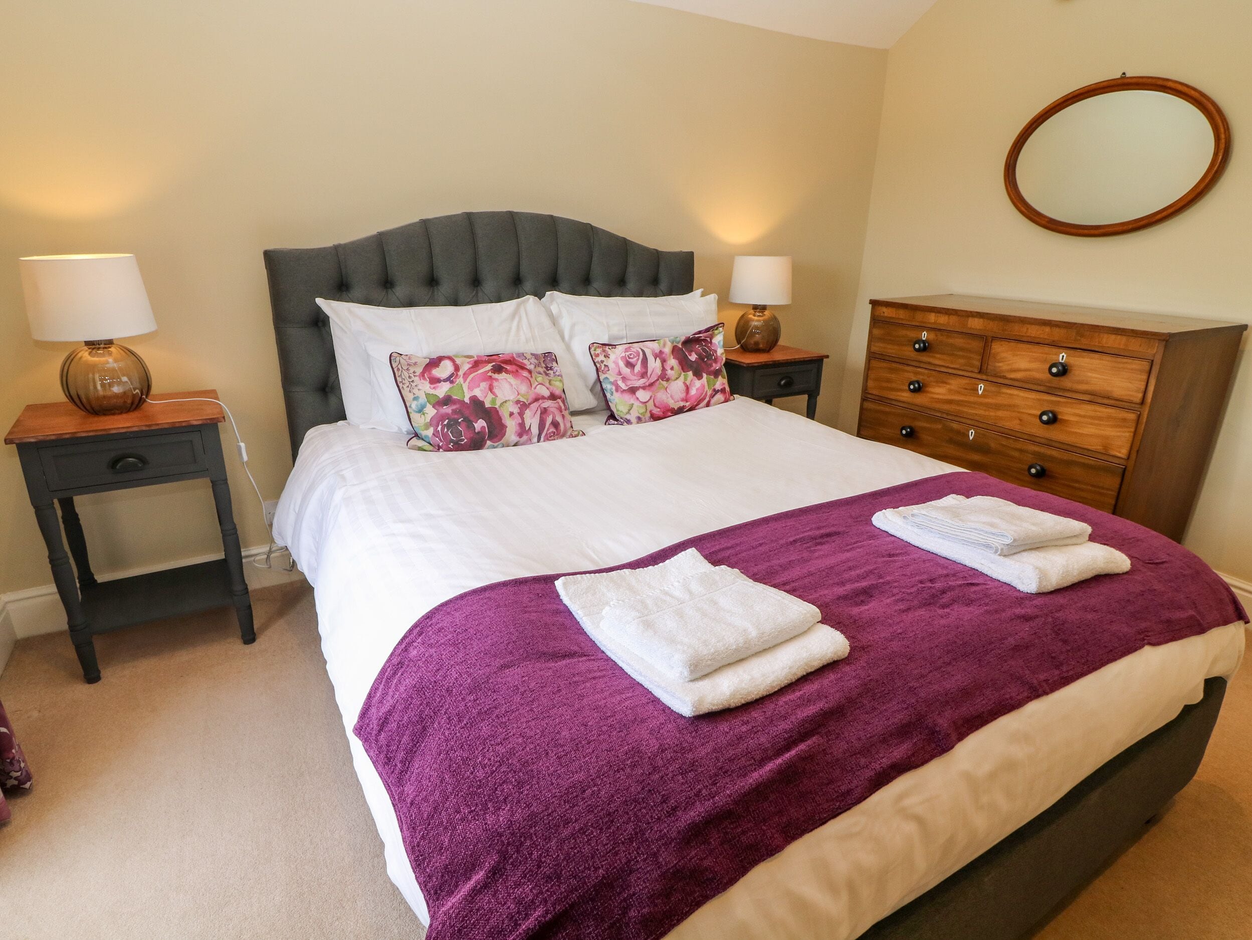 A neatly made bed with a purple blanket and two white towels, flanked by two bedside tables with lamps, a wooden dresser, and a round mirror on the wall.