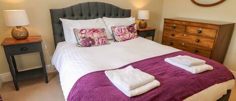 A neatly made bed with a purple blanket and two white towels, flanked by two bedside tables with lamps, a wooden dresser, and a round mirror on the wall.