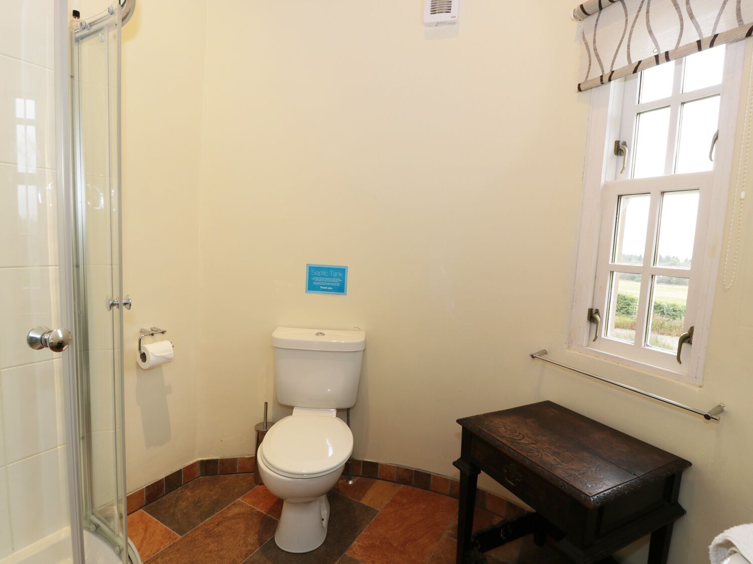 Une salle de bain avec des toilettes, une douche en verre, une petite table en bois et une fenêtre donnant sur un beau paysage.
