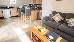 A modern kitchen with a dining area, a grey sofa with plaid cushions, and a wooden coffee table with a plant and a black folder. - The Wynnstead Annexe (Oswestry)
