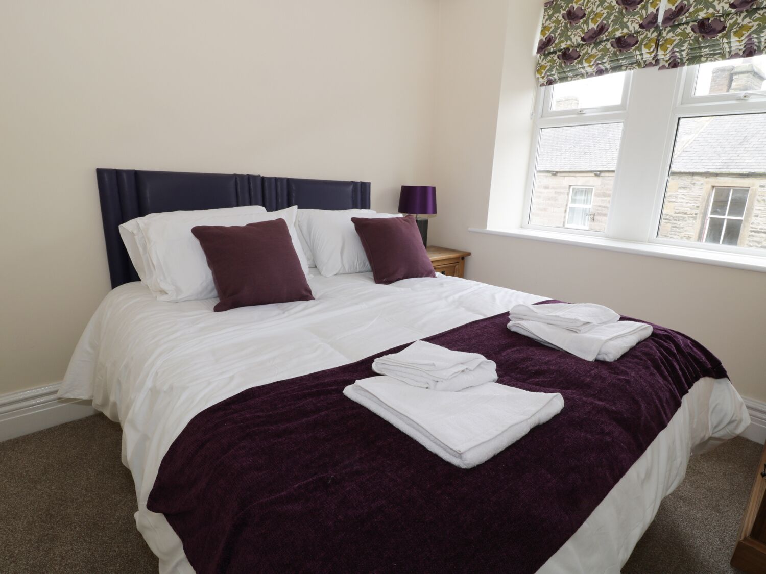 A neatly made bed with white linens and a purple throw blanket, a bedside table with a lamp, and a window with a floral curtain.