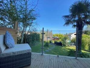 A wooden deck with a sofa and patterned pillows, overlooking a garden with a thatched-roof house and a palm tree. - Hearthstone East Cottage (Brixham)
