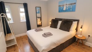 A neatly made bed with folded towels, a bedside lamp, a painting of sailboats, and a window with curtains. - South Beach at The Hideaway (Tenby)