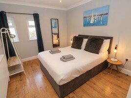 Una cama bien hecha con toallas dobladas, una lámpara de noche, un cuadro de veleros y una ventana con cortinas.