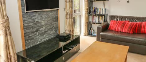 A living room with a flat-screen TV mounted on a stone wall, a bookshelf, a leather sofa with red cushions, and a wooden coffee table.
