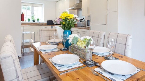 A modern dining area with a wooden table set for four, white plates, and a vase with yellow flowers. - Tus Nua Cottage (Minehead)