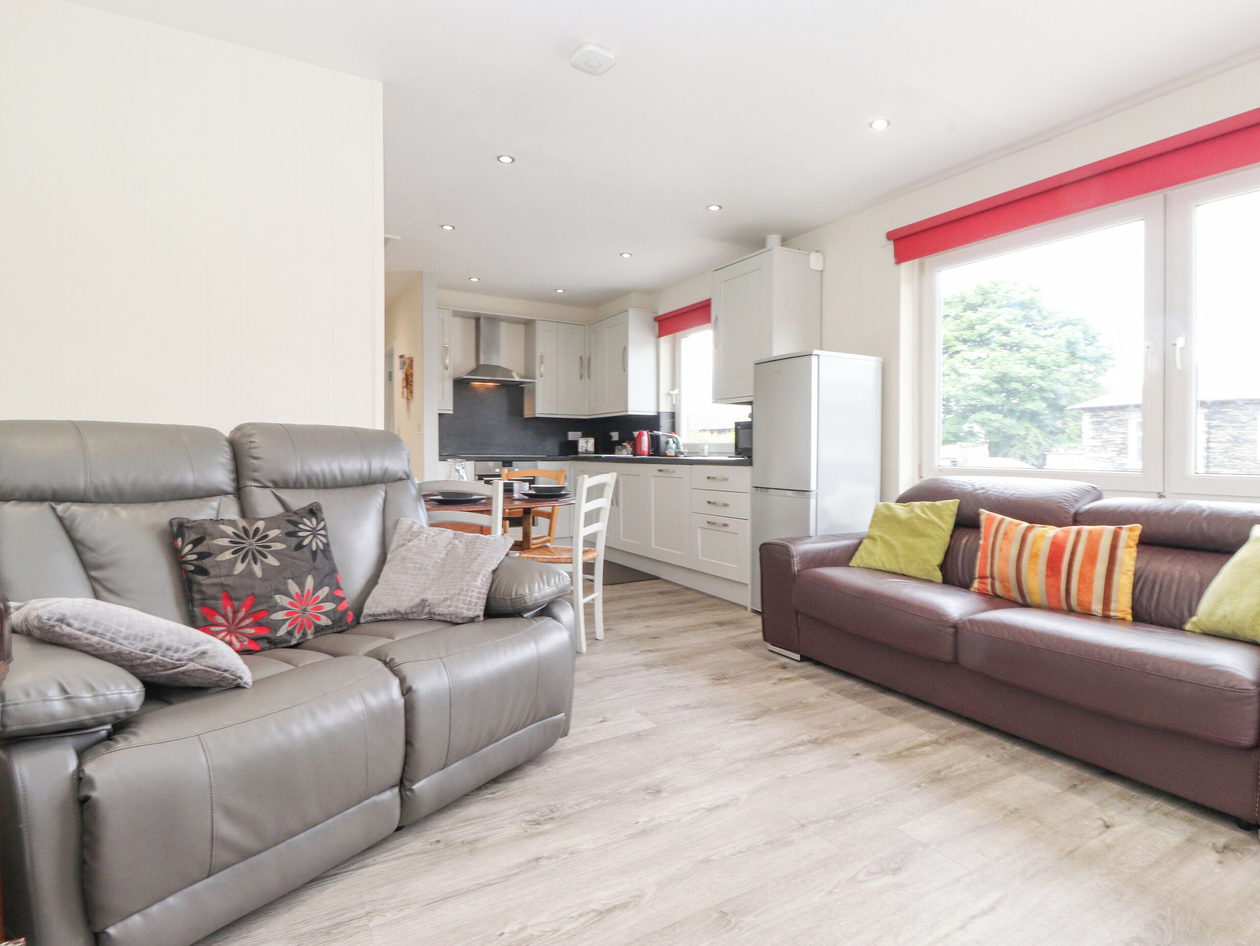 Ein modernes Wohnzimmer mit einem grauen Sofa und einem braunen Sofa, beide mit Kissen. Im Hintergrund steht ein Esstisch mit Stühlen. Die Wände sind weiß, der Boden ist aus Holz, und ein Fenster mit roten Vorhängen ist zu sehen.