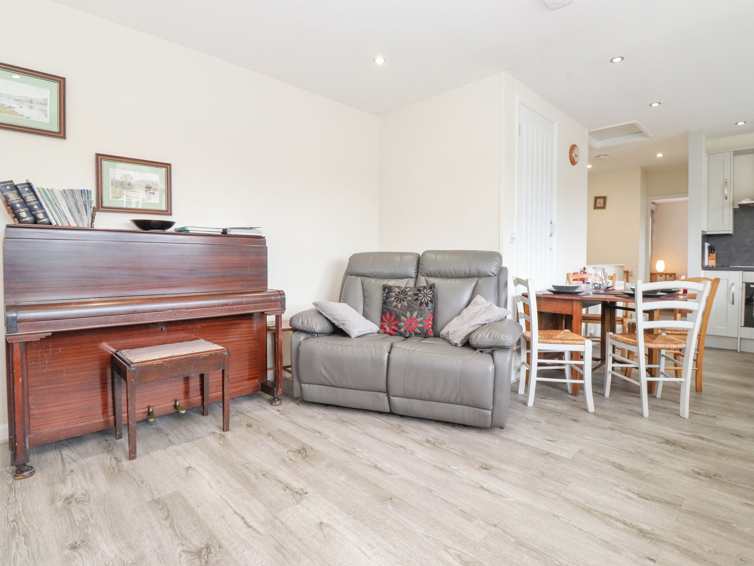 A living room with a piano, a grey sofa with cushions, a dining table with chairs, and a kitchen area in the background.