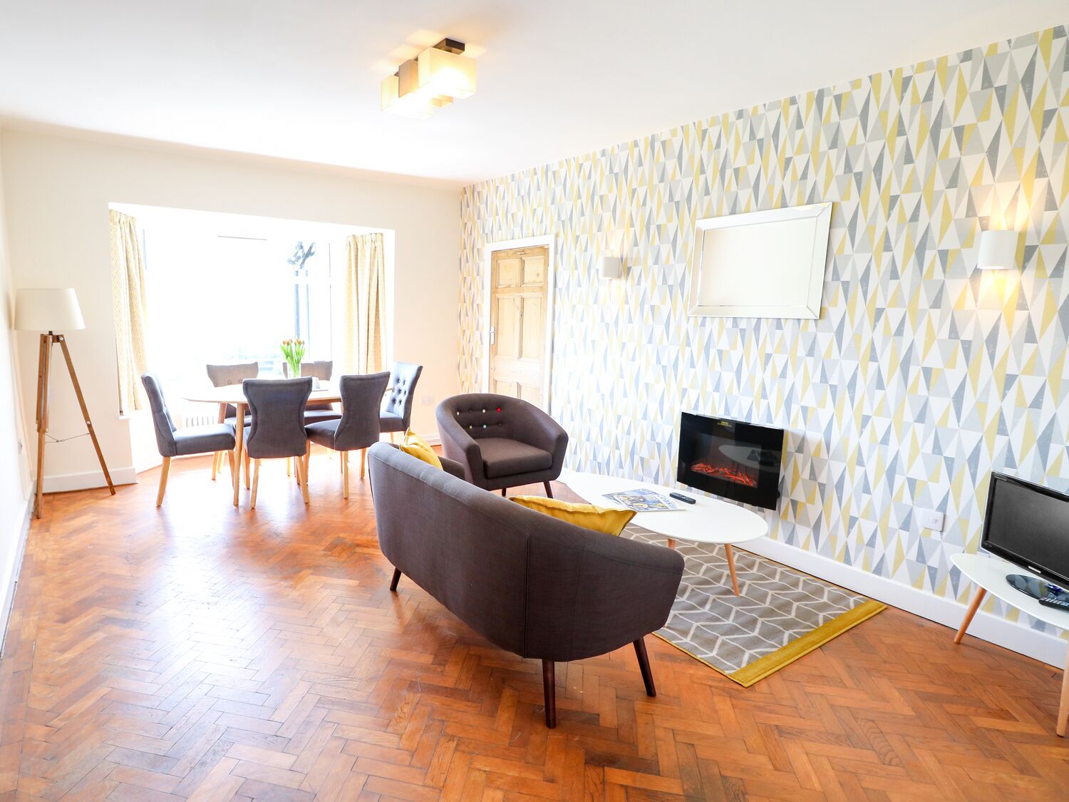 A modern living room with a herringbone wood floor, grey sofas, a flat-screen TV, and a patterned wallpaper.