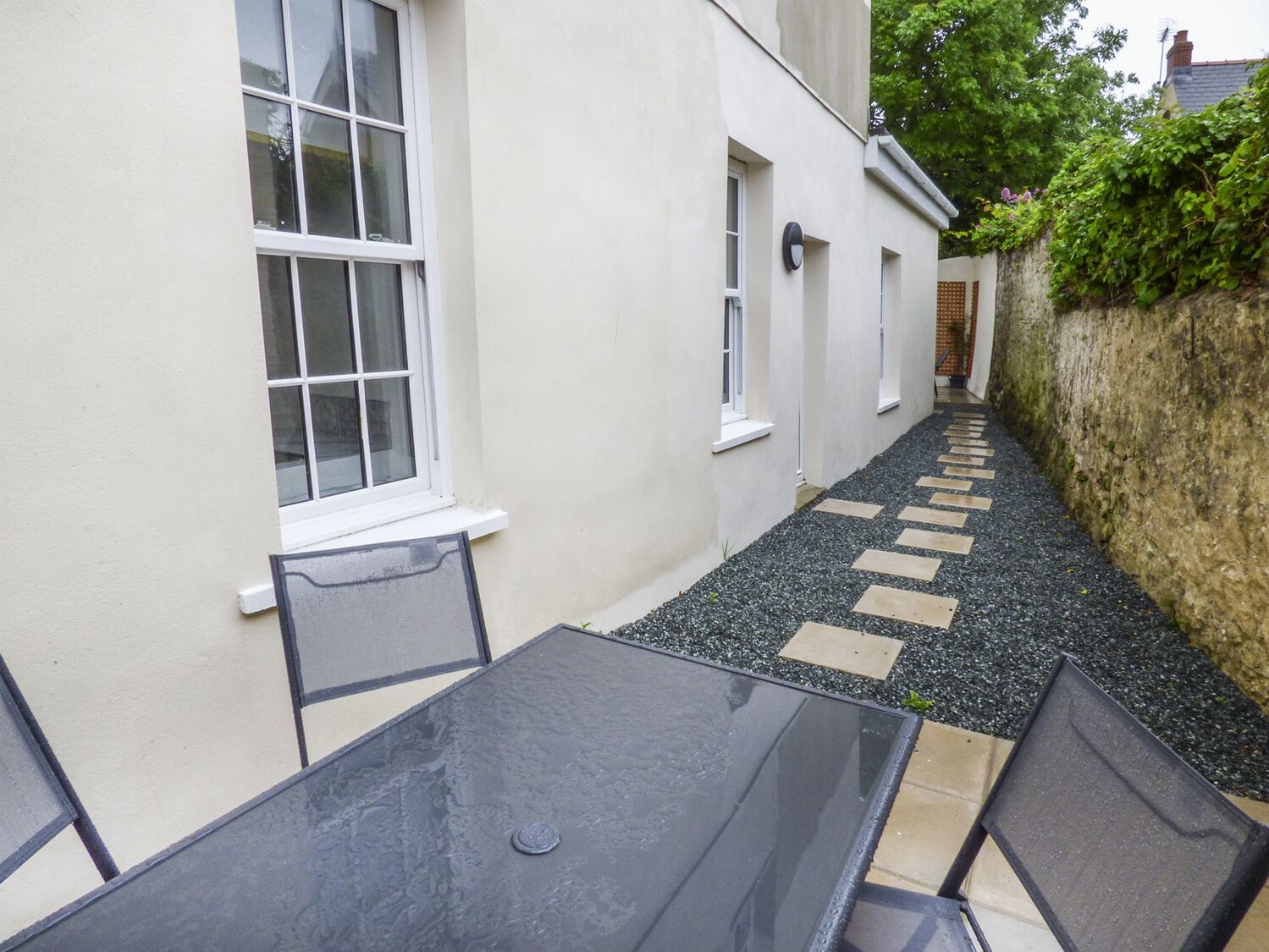 Un étroit couloir extérieur avec un chemin dallé, une table en verre et des chaises.