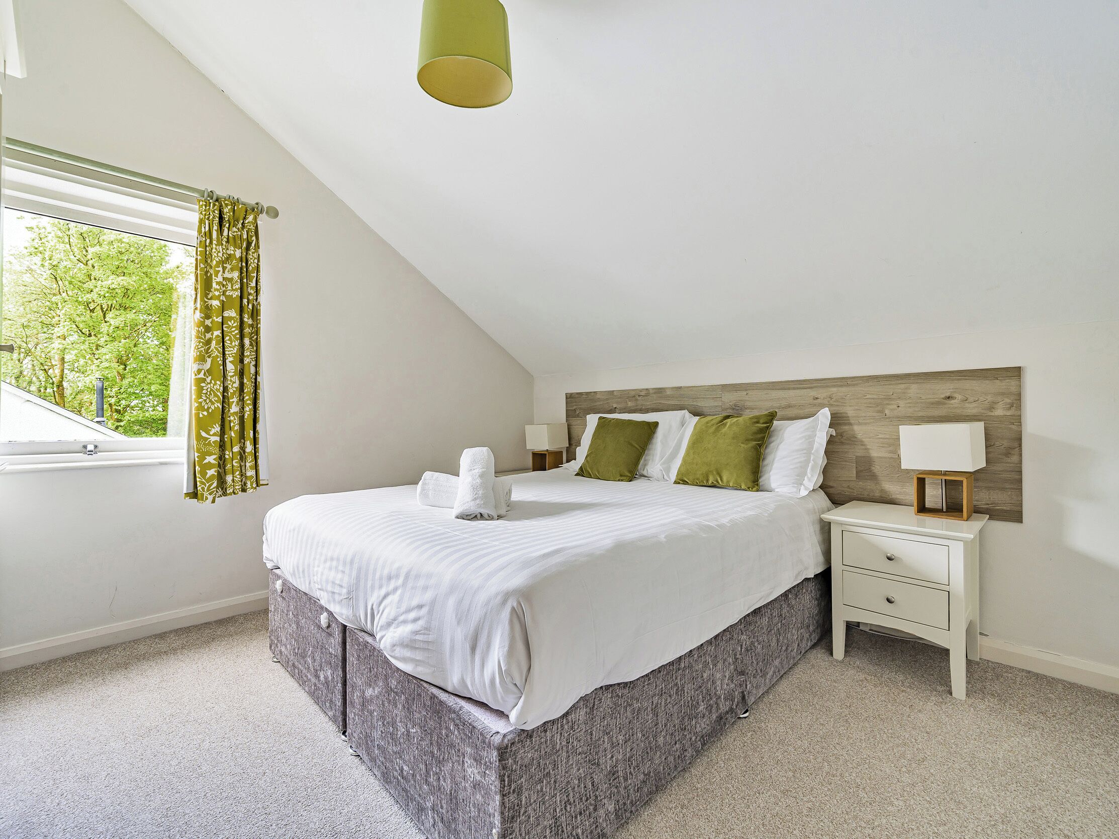 A bedroom with a sloped ceiling, a bed with white bedding, two green pillows, a wooden headboard, a bedside table with a lamp, and a window with curtains.