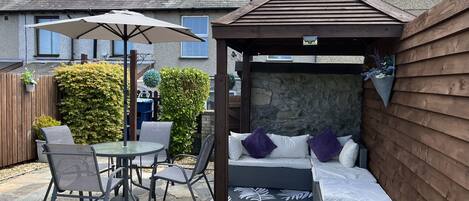 A patio area with a table and chairs, a covered seating area with cushions, and a stone floor.