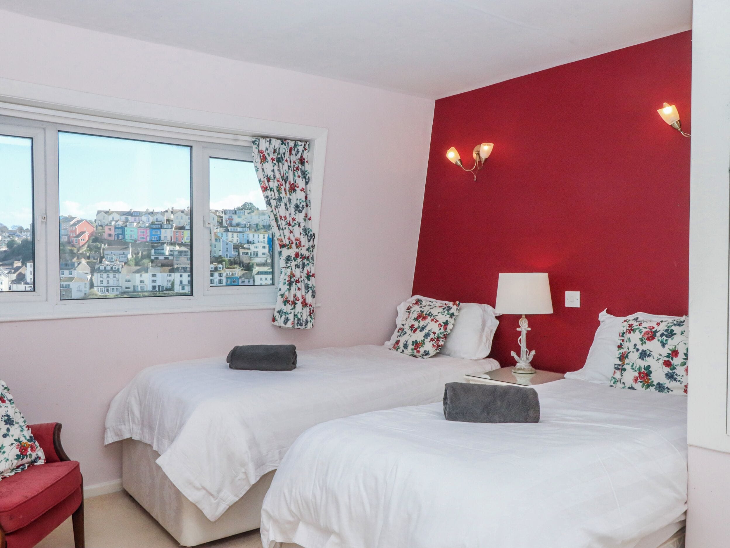 Ein Hotelzimmer mit zwei Betten, einem Fenster mit floralen Vorhängen, einer roten Akzentwand und Blick auf Gebäude.