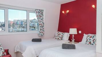 A hotel room with two beds, a window with floral curtains, a red accent wall, and a view of buildings outside.