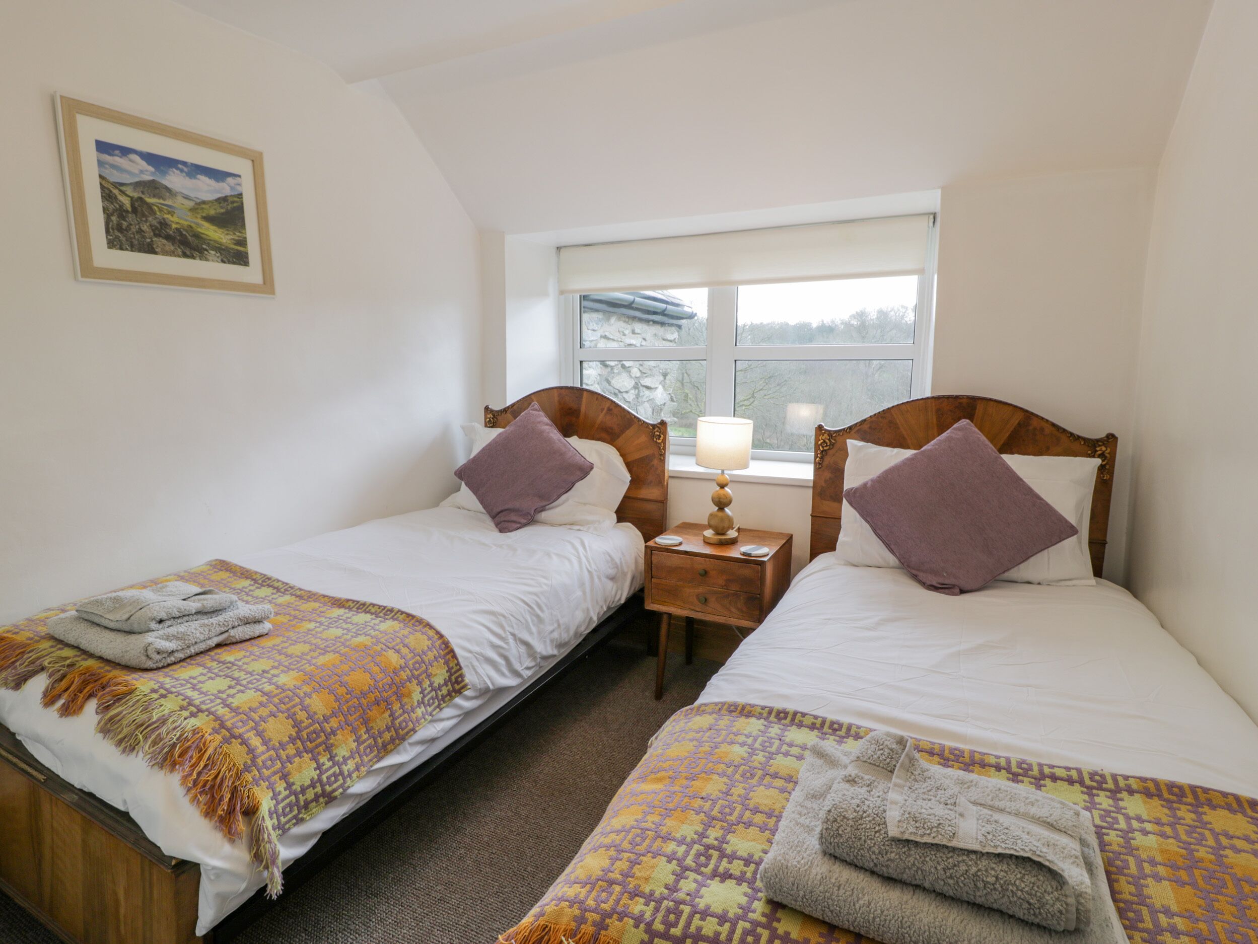 A room with two single beds, a nightstand with a lamp, a window with a view of trees, and a framed picture on the wall.