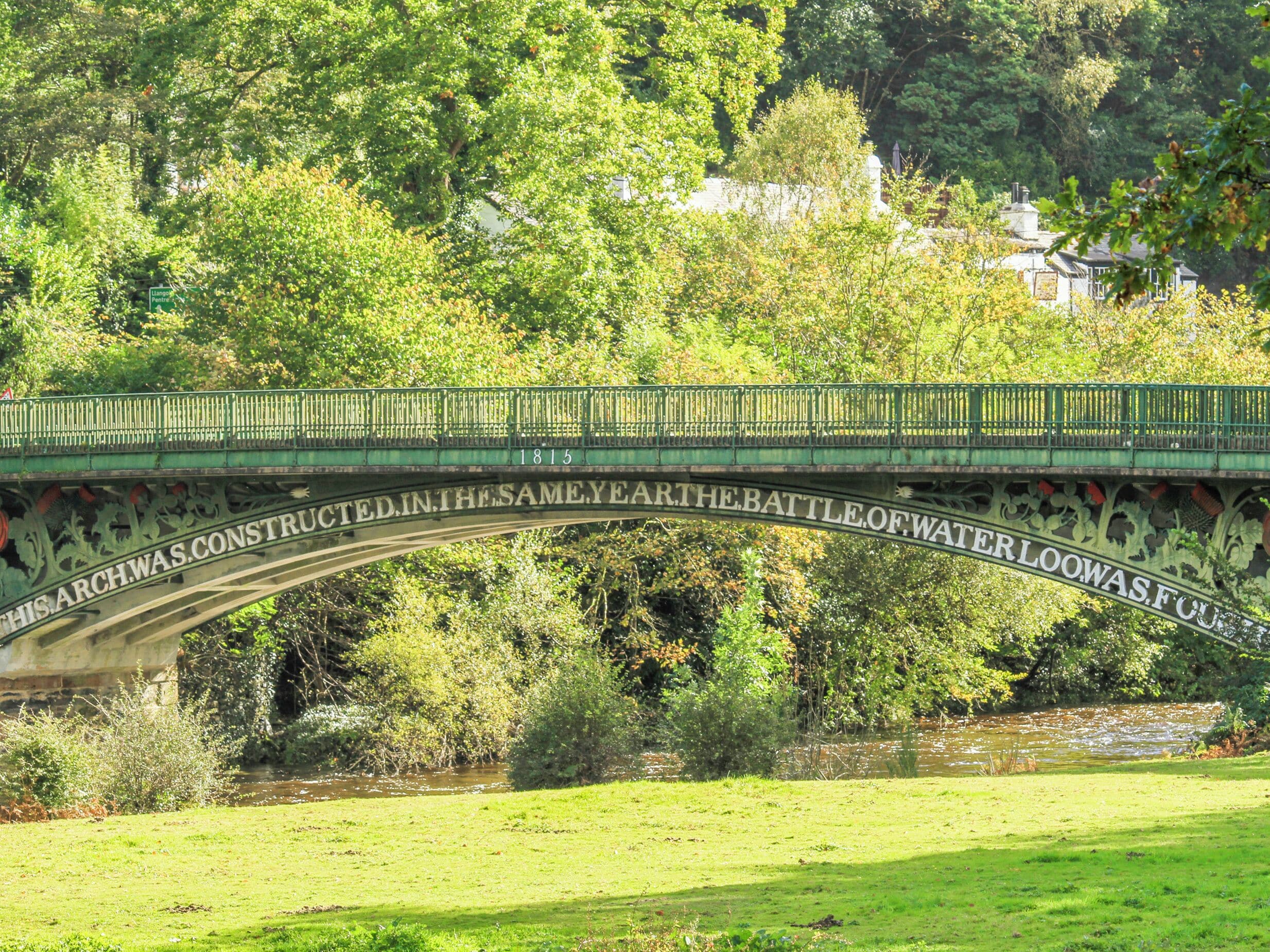 A green bridge with historical inscriptions, surrounded by lush greenery and a calm river.