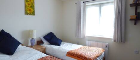 A room with two single beds, a window with curtains, a radiator, and a wall-mounted shelf with books and a teddy bear.