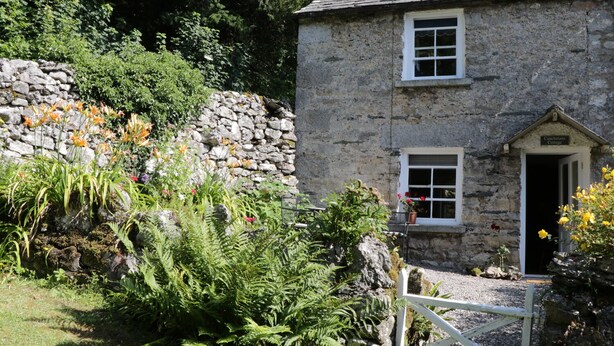 A stone cottage with a white door and window shutters, surrounded by lush greenery and flowering plants. - Coachmans Cottage (Grange-over-Sands)