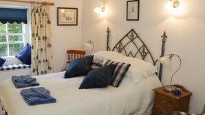 A neatly made bed with pillows and a folded towel, a bedside lamp, a wooden nightstand, and a chair with a striped cushion. - Coachmans Cottage (Grange-over-Sands)
