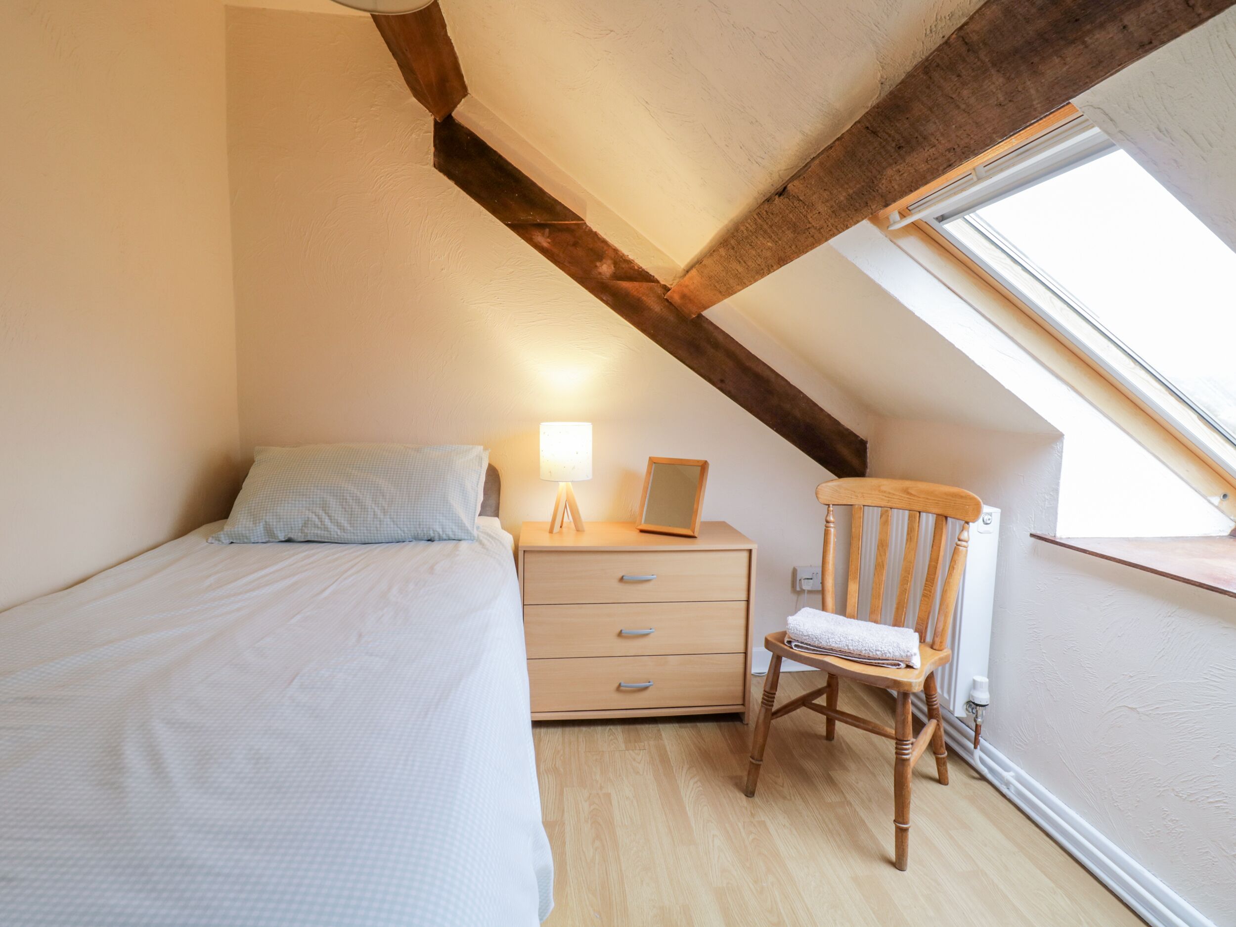 A bedroom with a single bed, a wooden nightstand with a lamp, a chair, and a window.