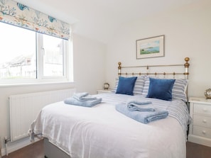 A neatly made bed with striped bedding and blue pillows, a window with a view of a building, and a framed picture on the wall. - Gull Cottage (Padstow)