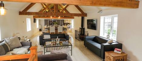 A spacious living area with a high ceiling and exposed wooden beams, a dining area, a fireplace, and a large window providing natural light.