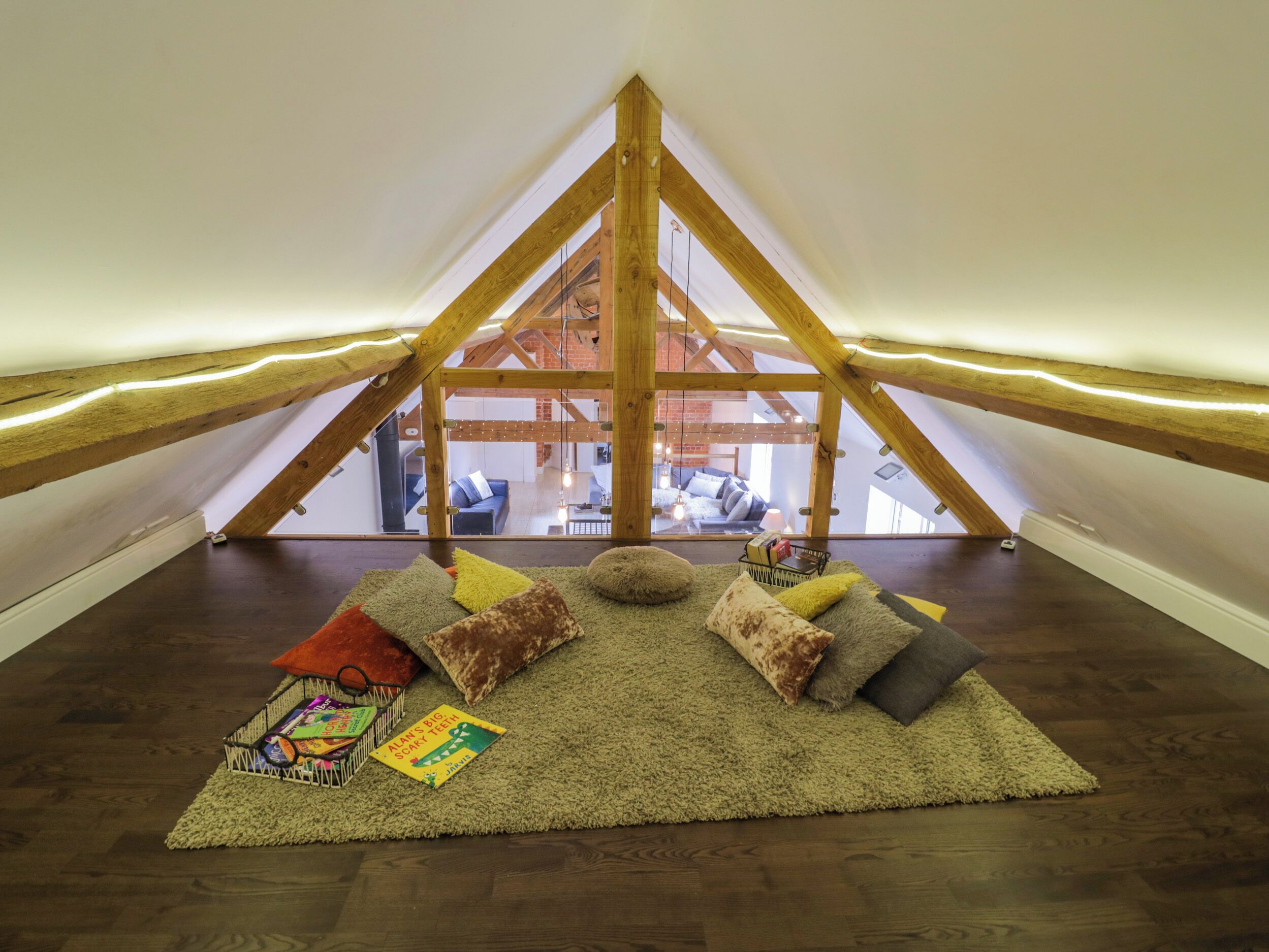 Un dormitorio en ático con techo inclinado, vigas de madera, vista a una sala de estar y un conjunto de almohadones y alfombra acogedor.