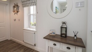 A room with a radiator, a window with striped valance, a round mirror, a small wooden console table with drawers, and a wall-mounted decorative item.