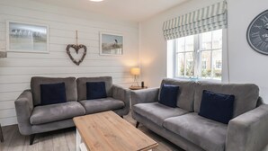 A living room with two grey sofas, a wooden coffee table, a hanging light fixture, a wall clock, and a window with striped curtains.