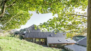 A stone house with a sloped roof, surrounded by lush greenery and a hill.