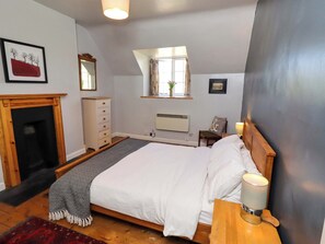 A bedroom with a bed, a dresser, a mirror, a window, a fireplace, and a nightstand with a lamp. - Gamekeepers Cottage (Alnwick)