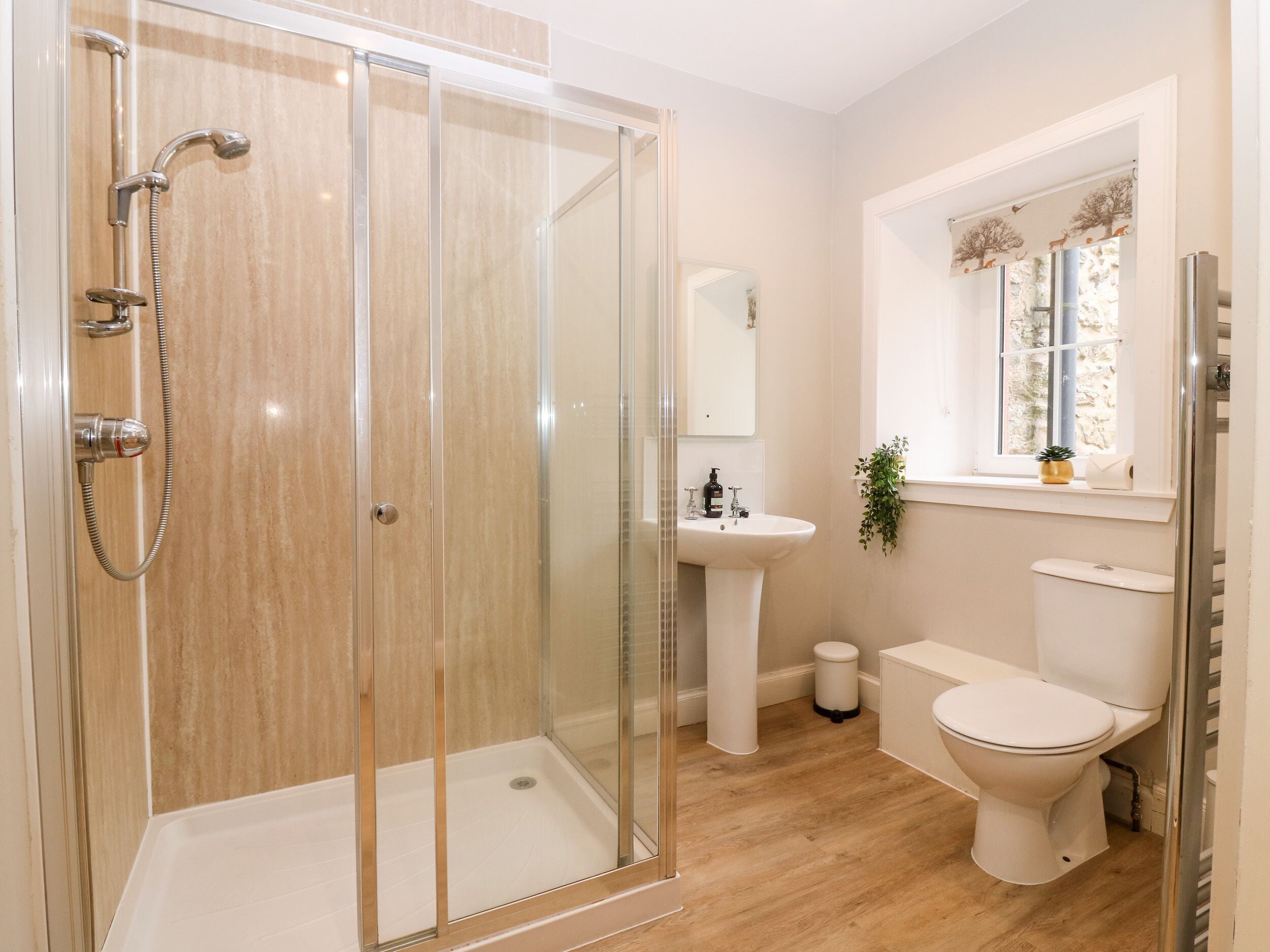 Een moderne badkamer met een douchecabine van glas, een wastafel op een voetstuk, een toilet en een raam met een plant.