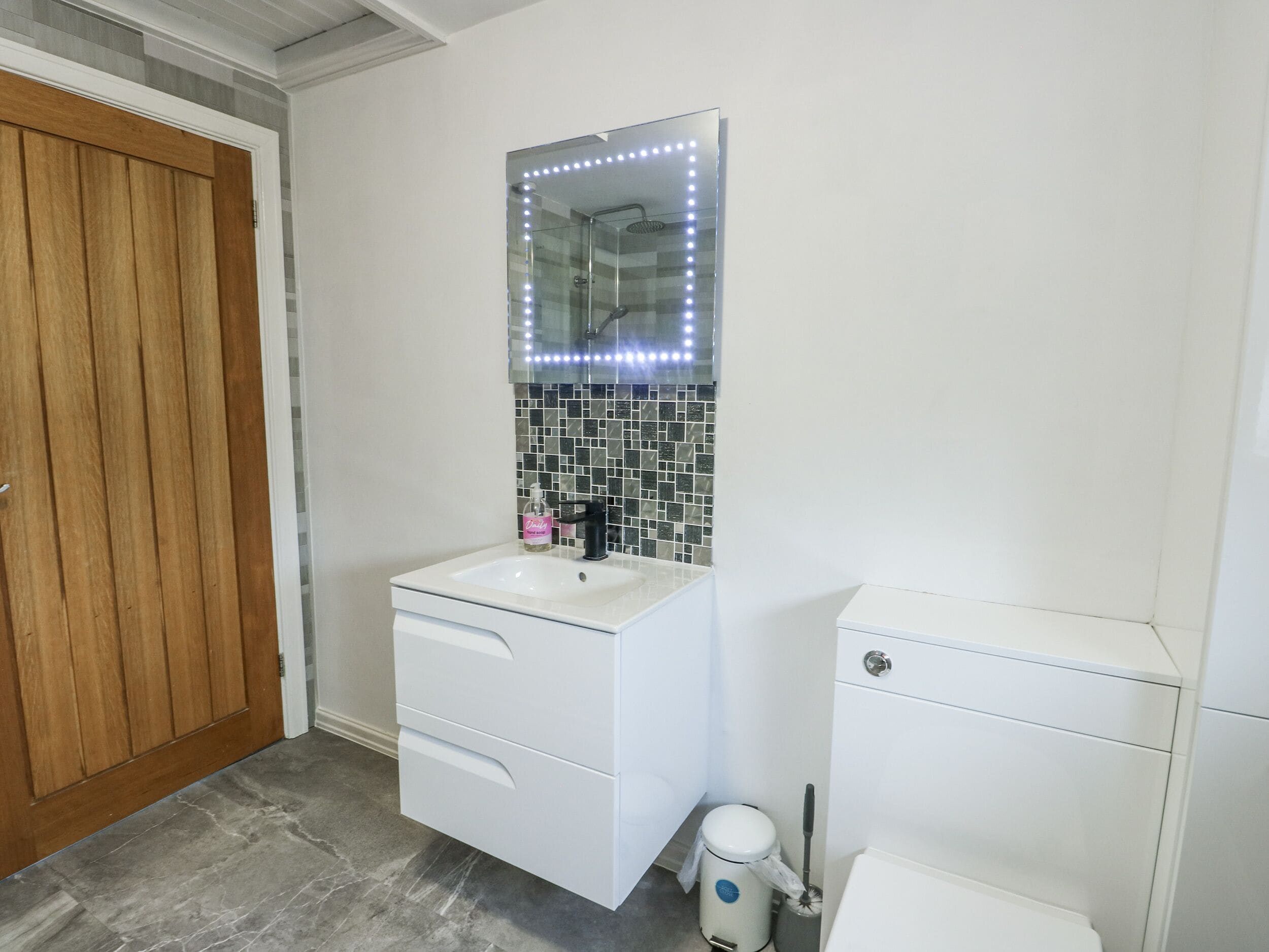 Une salle de bain avec un lavabo blanc, un miroir avec éclairage à DEL, un mur recouvert de carreaux, une porte en bois et des toilettes.