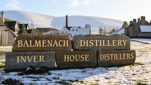 A sign with 'BALMENACH DISTILLERY HOUSE DISTILLERS' in front of a building with a snowy landscape and mountains in the background. - Corriemhor Beag (Grantown-on-Spey)