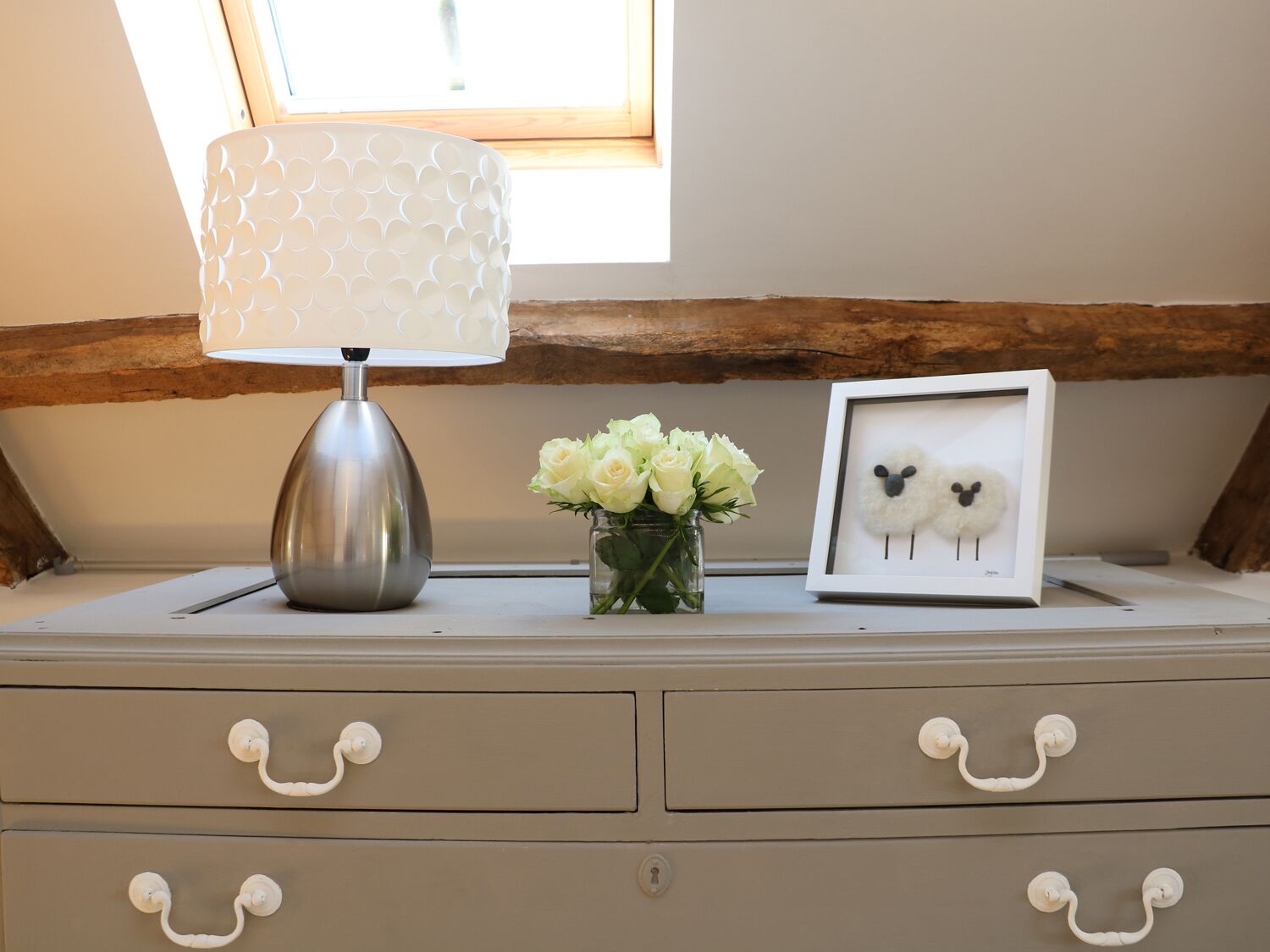 A bedside table with a lamp, a framed picture of sheep, and a vase of flowers.