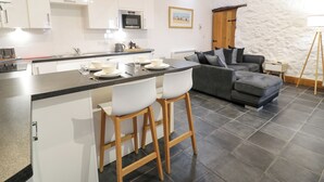 A modern kitchen with white cabinets, a central island with bar stools, a grey sofa, and a stone wall. - Cornel Glyd (Talybont)