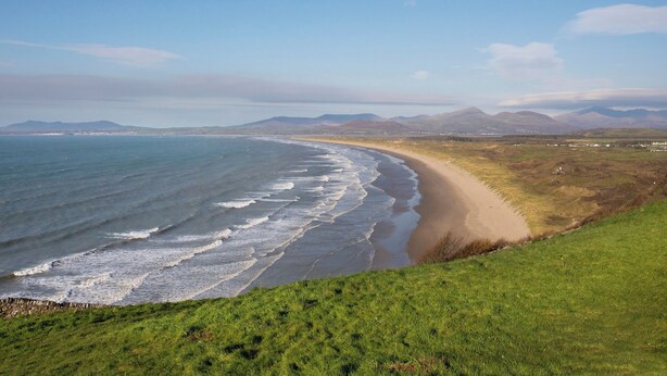 Un paysage côtier avec une plage, des zones herbeuses et des montagnes à l’horizon.