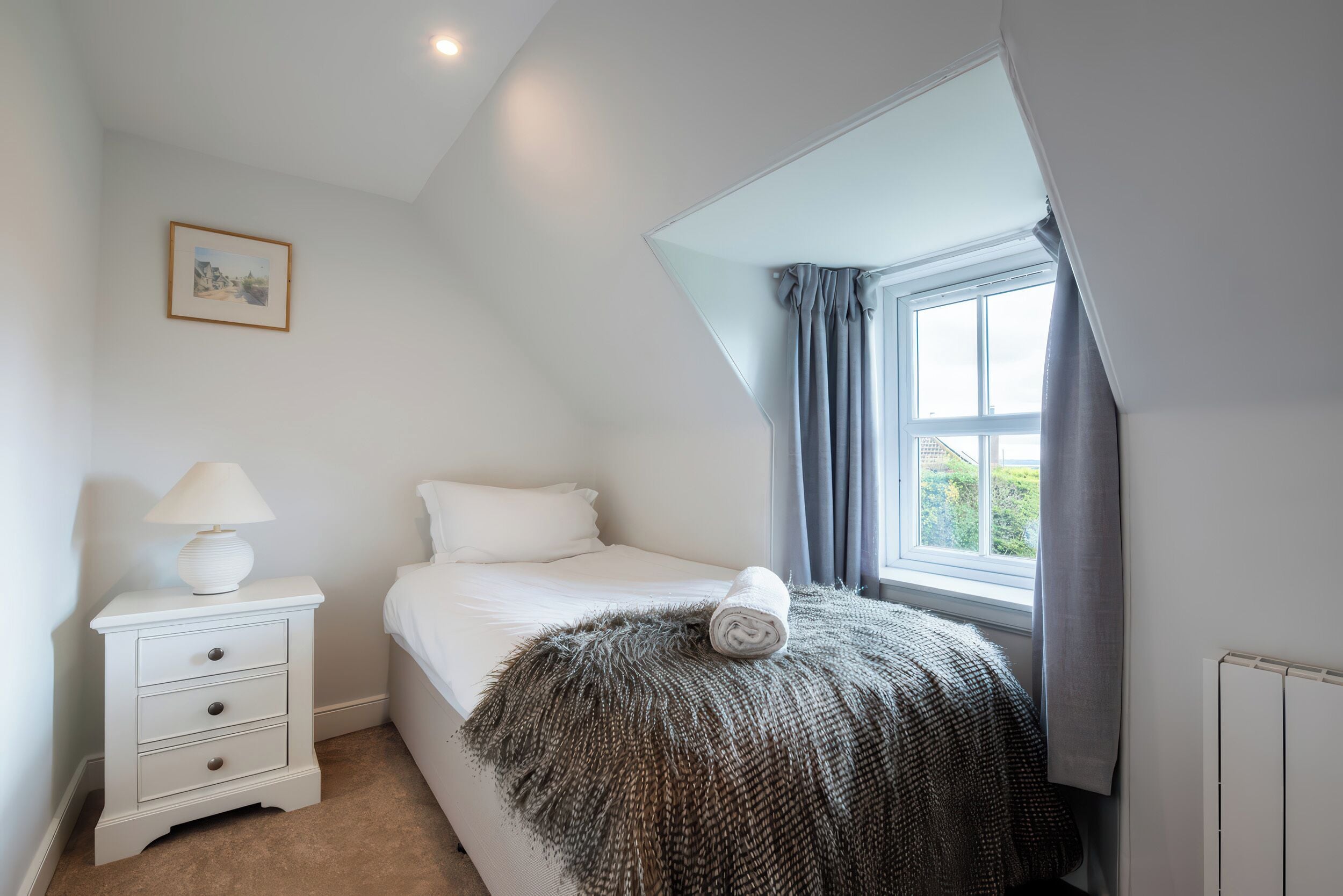 A bedroom with a bed, a nightstand with a lamp, a window with curtains, and a framed picture on the wall.