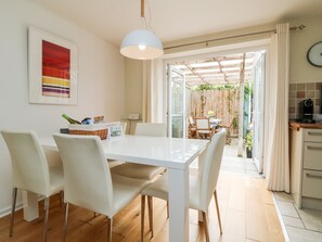 Ein moderner Essbereich mit einem weißen Tisch und Stühlen, einer Hängeleuchte und Blick auf eine Terrasse mit Holzfußboden und Pflanzen.