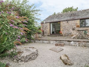 Un bâtiment en pierre avec une allée gravillonnée, une table en bois et un pot de fleurs.