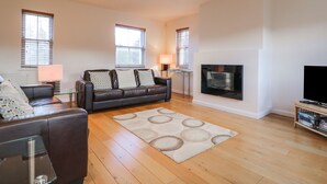 A spacious living room with a leather sofa, a flat-screen TV mounted on the wall, and a wooden floor. - Wrenville (Keswick)