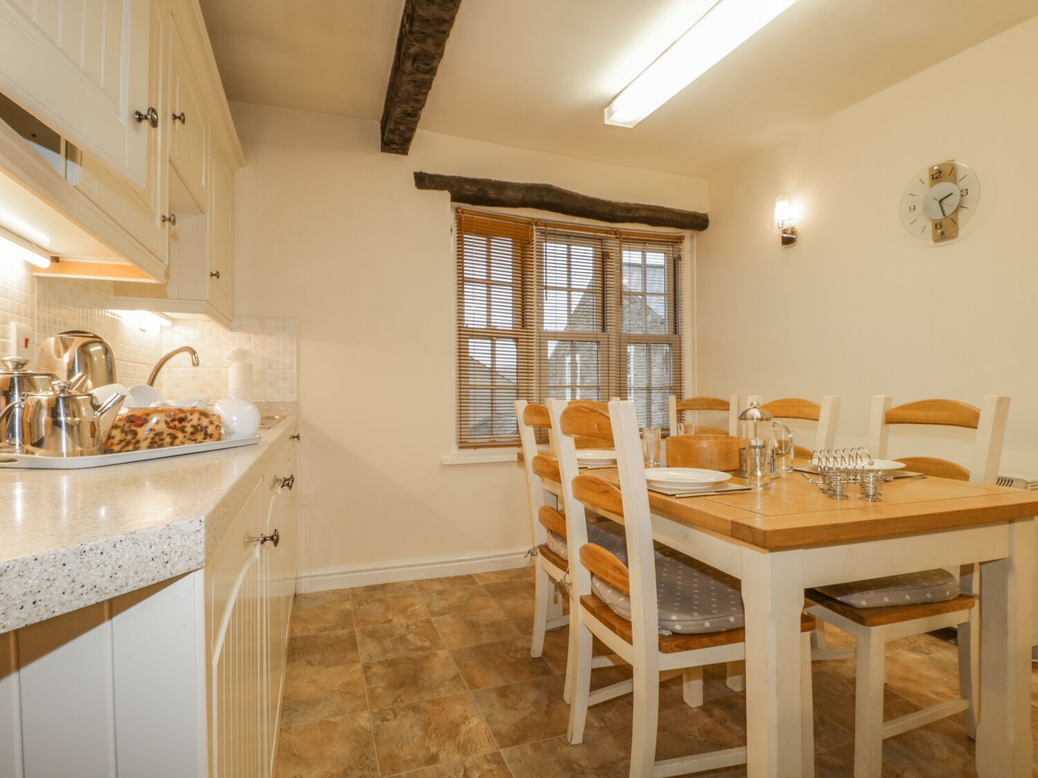 A dining area with a wooden table and chairs, a kitchen with white cabinets and a granite countertop, and a window with blinds.