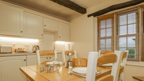 A dining area with a wooden table and chairs, a window with blinds, and white cabinetry. - Ale Cottage (Wigton)