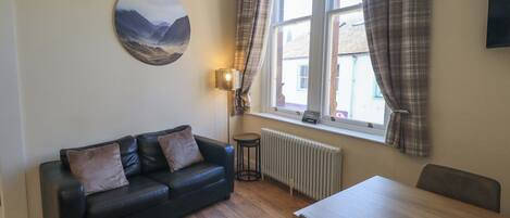 A living room with a black leather sofa, a round wall art, a standing lamp, a dining table, and a window with plaid curtains.