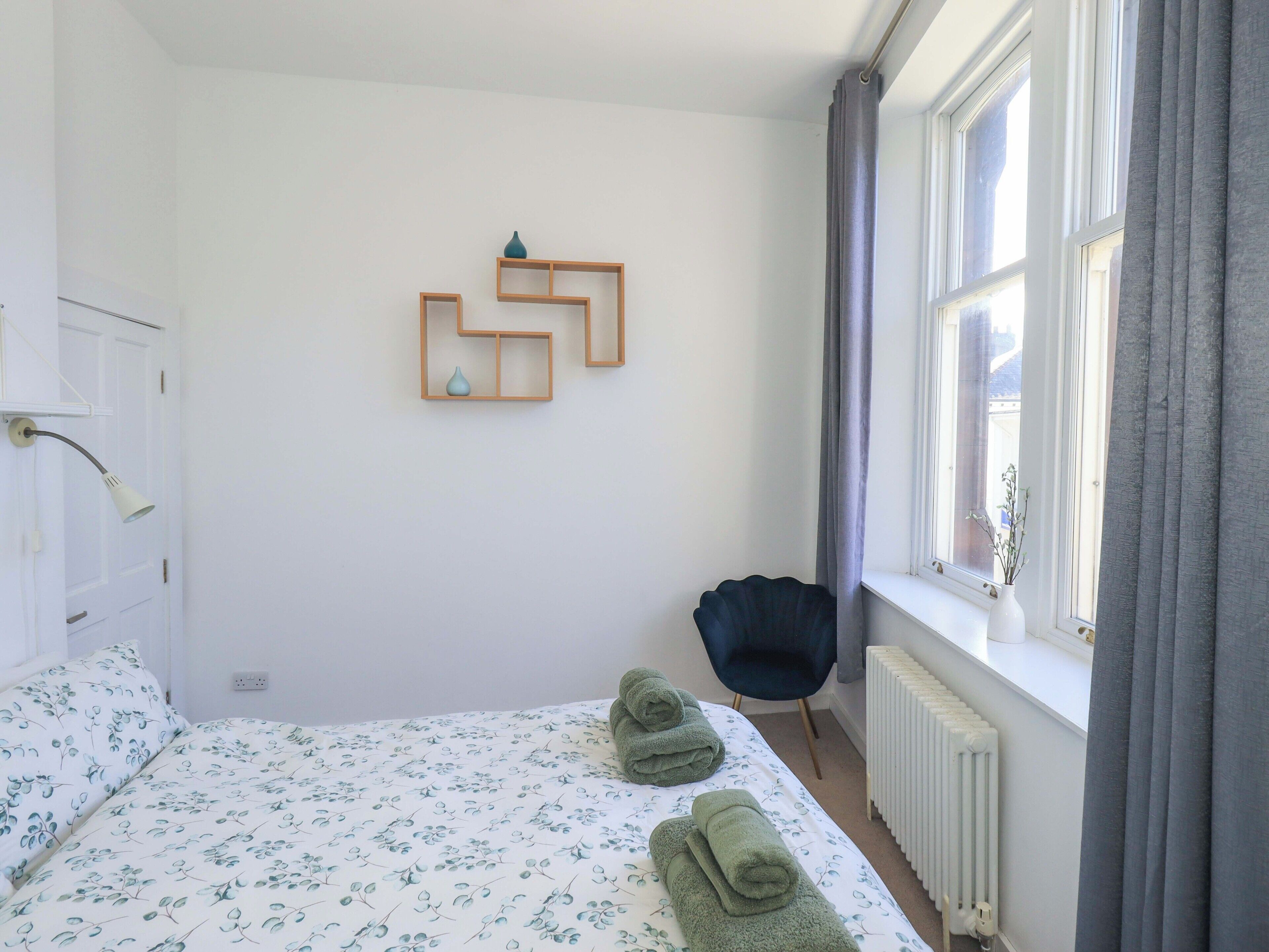Un dormitorio con una cama, una silla, un radiador y una ventana con cortinas.