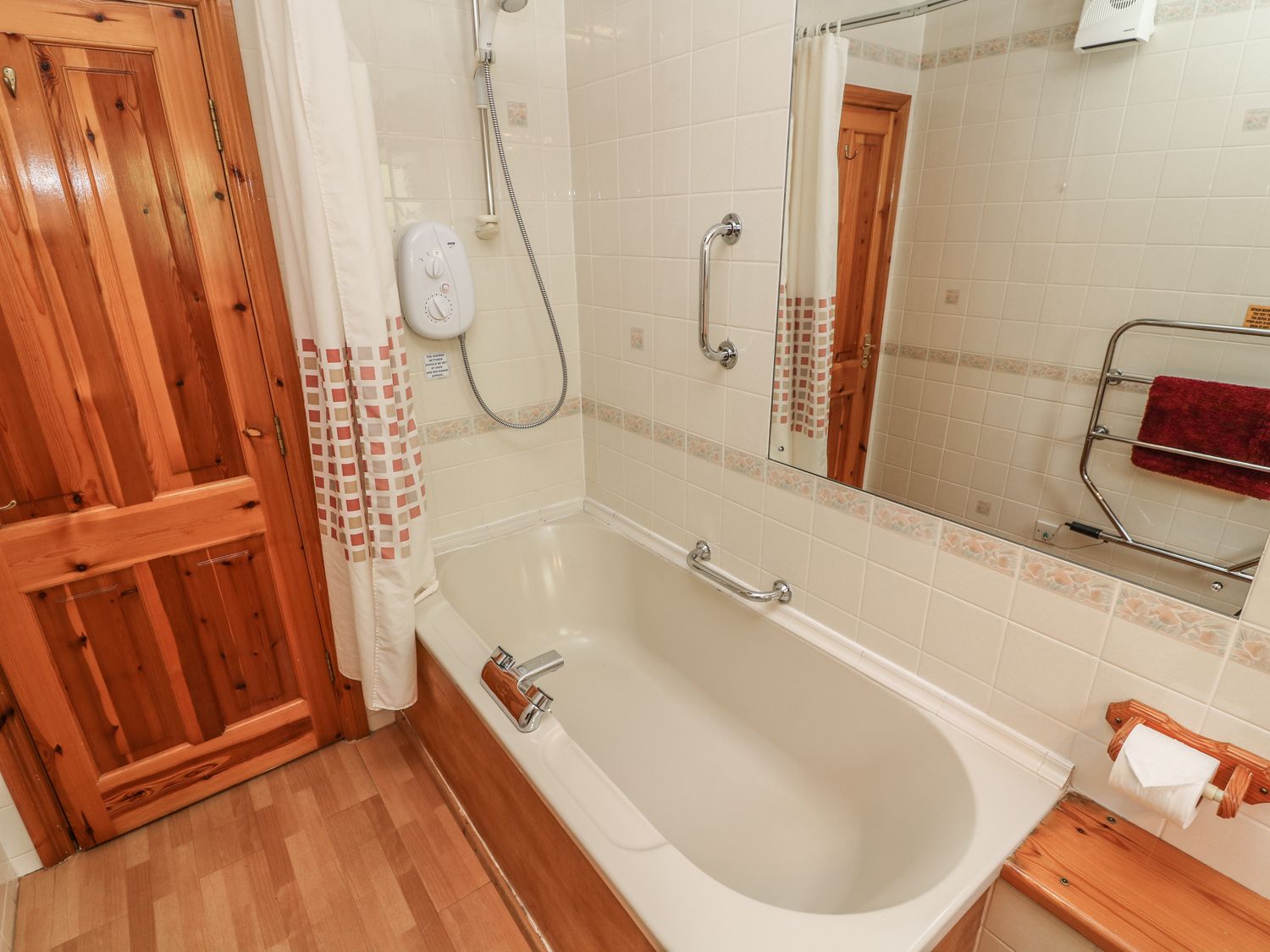 A bathroom with a wooden door, a white bathtub, a shower, a handrail, a towel rack, and a mirror.