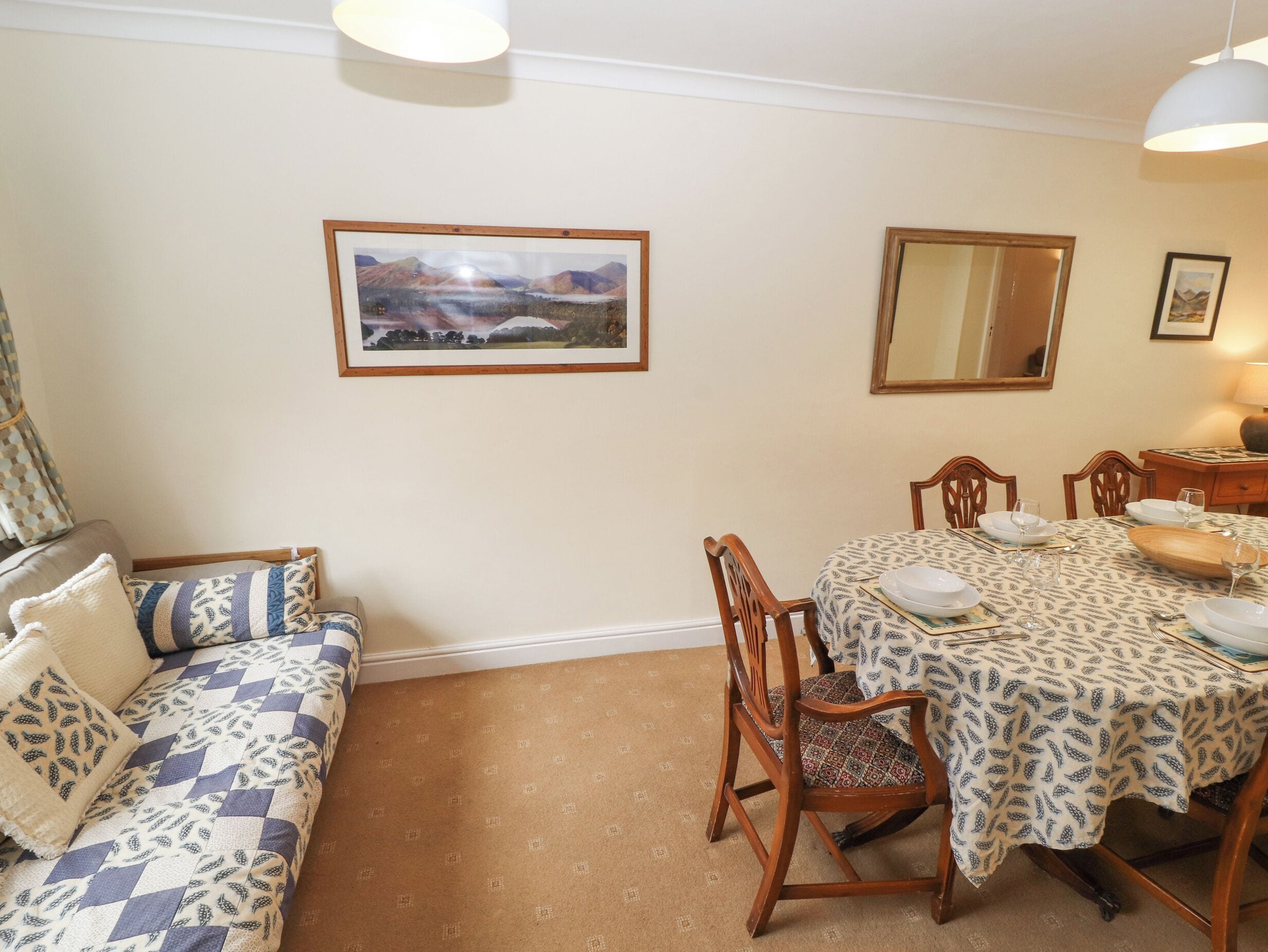 A dining area with a table set for four, a sofa with patterned cushions, and a framed landscape painting on the wall.