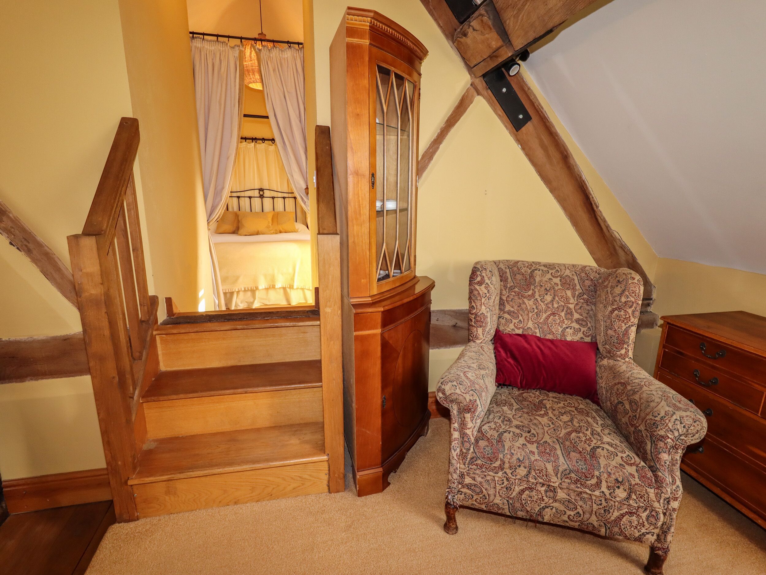 A bedroom with a wooden staircase, a bed with white bedding, a wooden cabinet, and a patterned armchair.