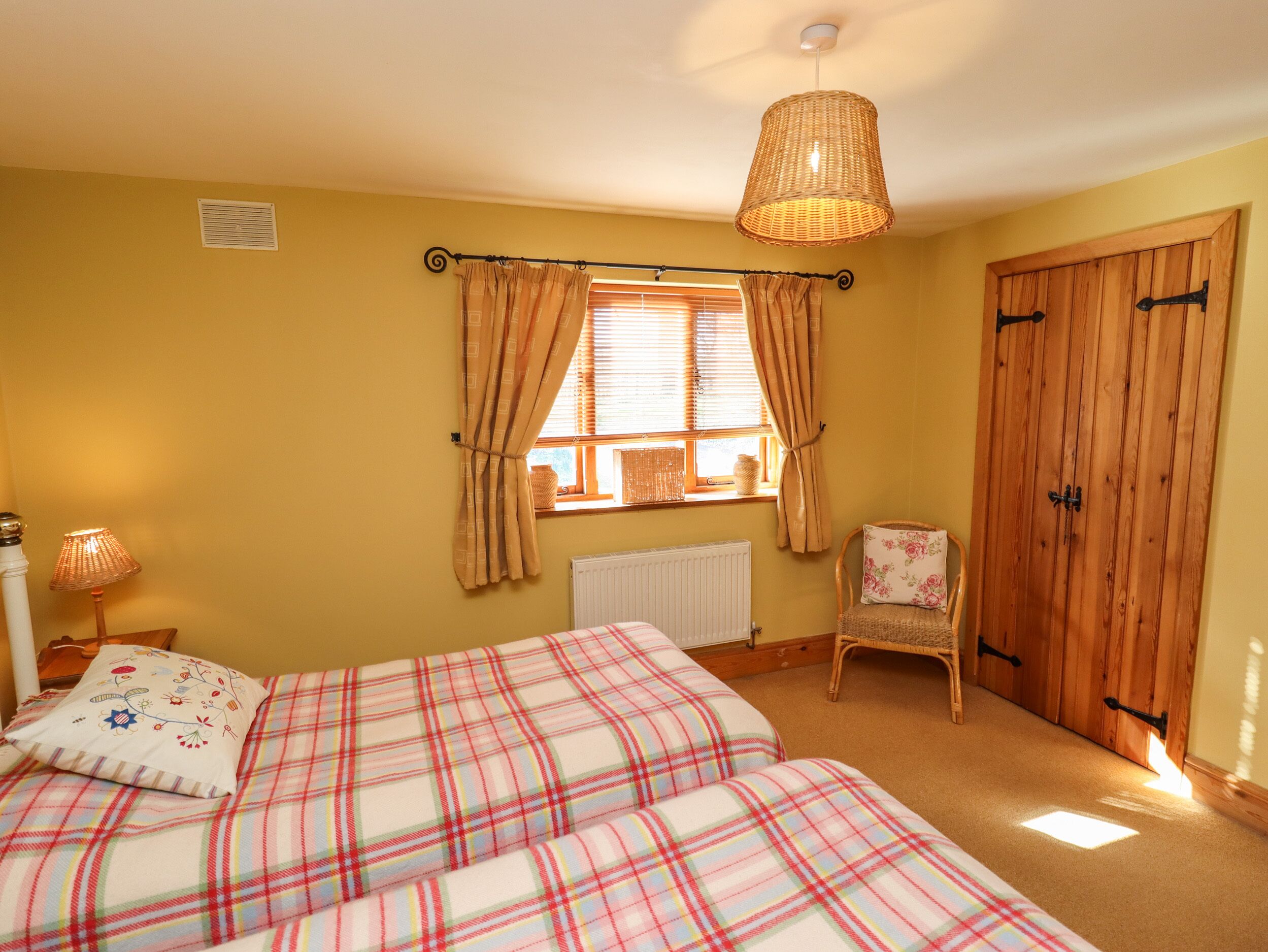 A bedroom with two beds, a window with curtains, a radiator, a wicker chair, and a wooden wardrobe.